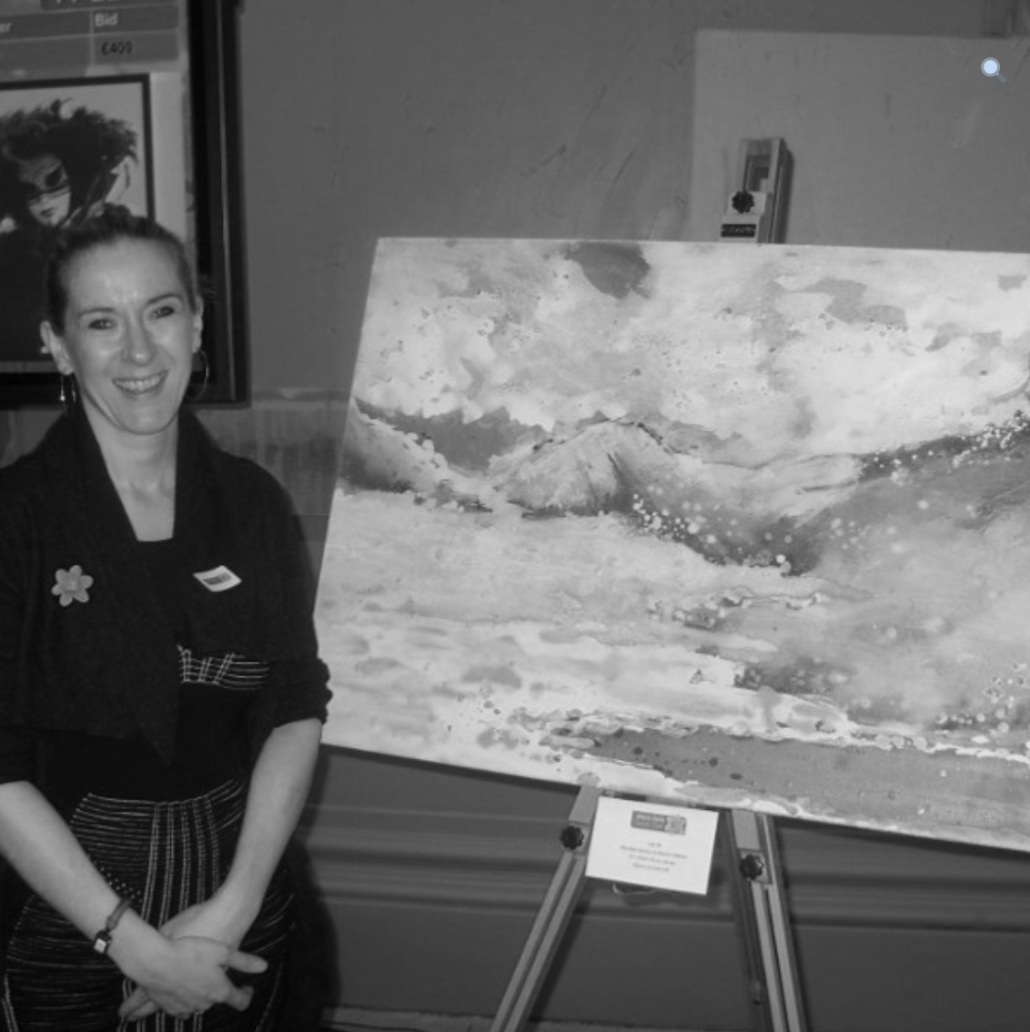Black and white photo of artist Sharon Withers standing by a landscape painting on an easel
