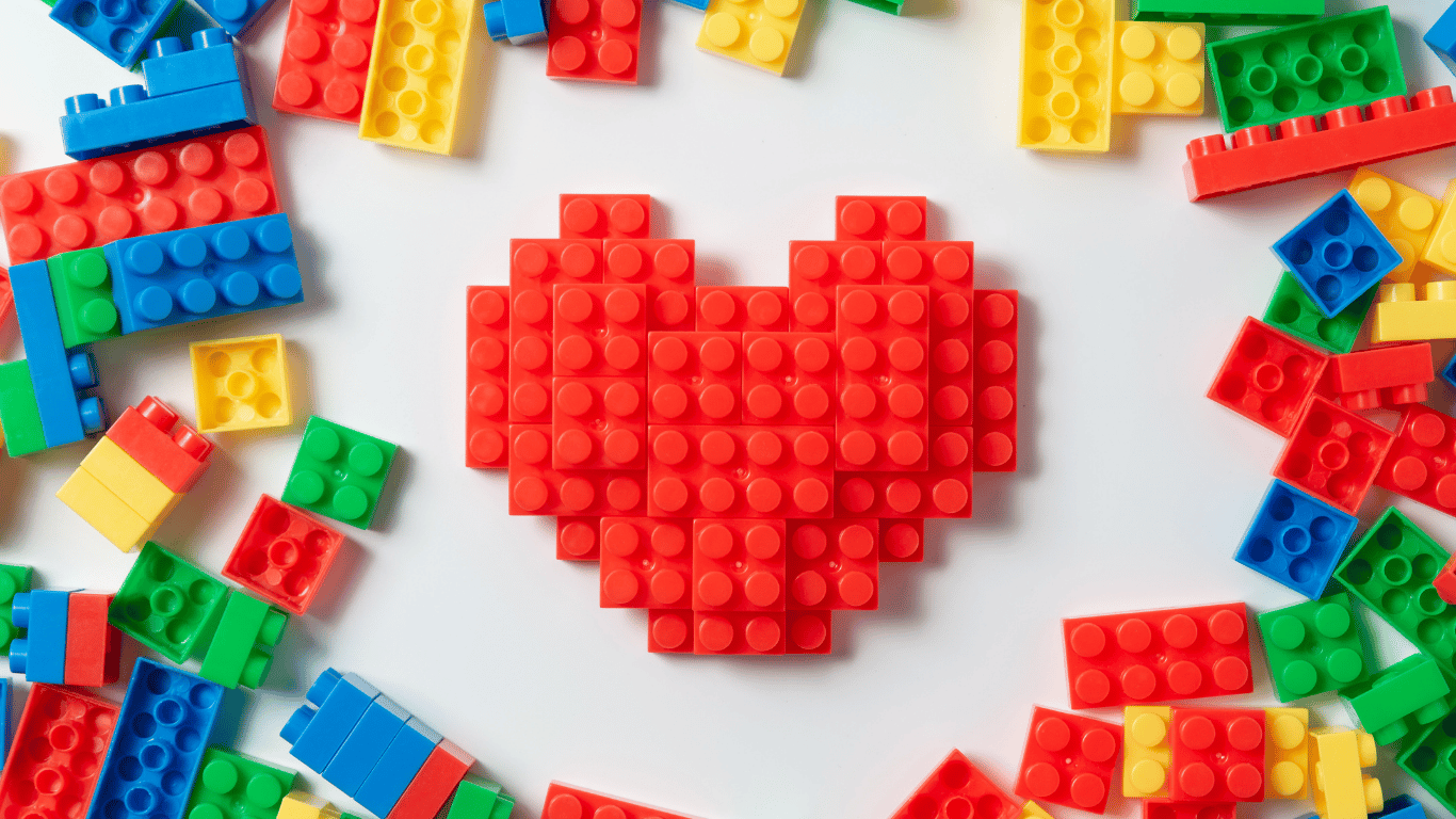 Photo of a red heart made of LEGO pieces and colourful blue, yellow, green and red lego scattered around the outside of the heart on a white background