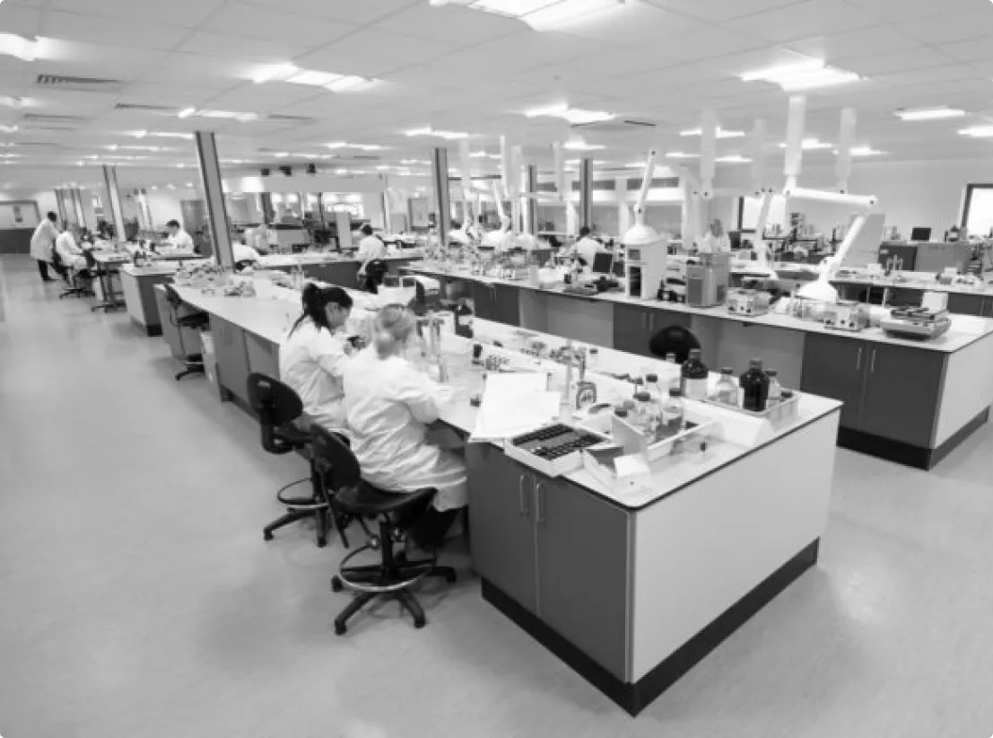 Wide view of a busy laboratory with scientists in white coats working at long benches filled with lab equipment and instruments.