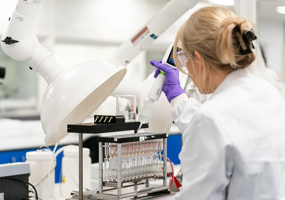 Scientist in a lab coat and purple gloves using a pipette to transfer liquid into test tubes in a laboratory setting.