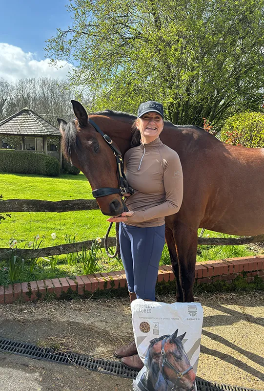 Smiling woman in riding gear standing next to a brown horse outdoors with a bag of horse feed in front.