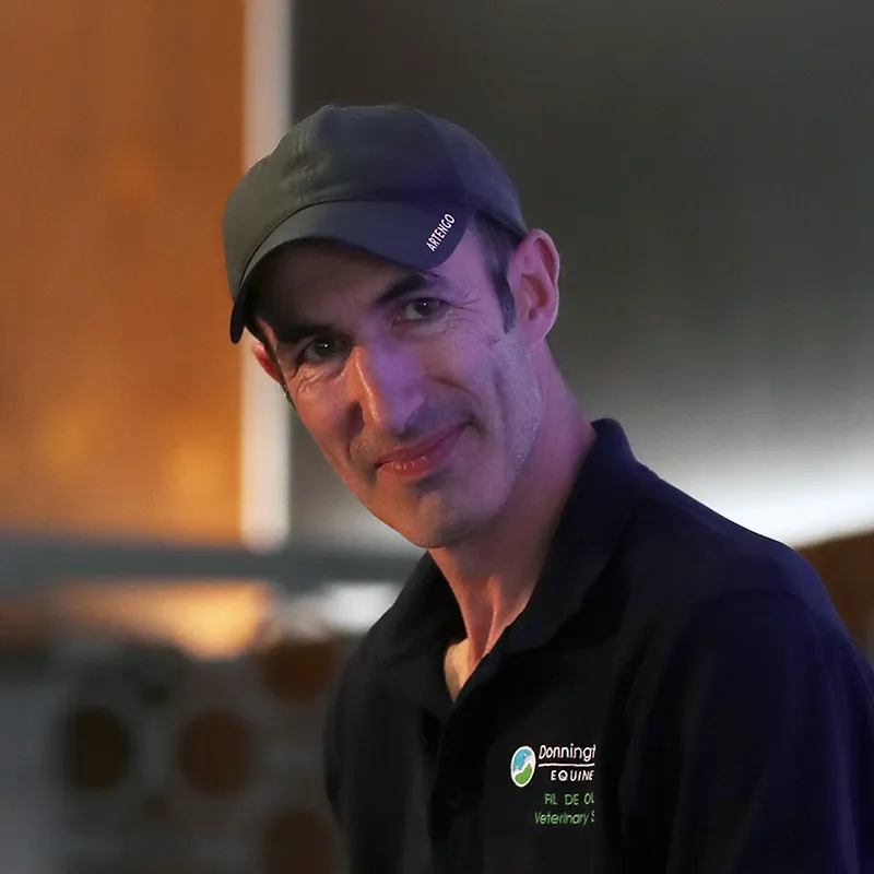 Man wearing a dark cap and black polo shirt with a veterinary service logo, smiling softly.