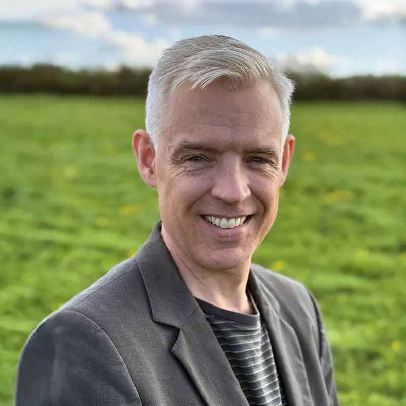 Smiling middle-aged man with gray hair wearing a suit jacket over a striped shirt in a green outdoor field.