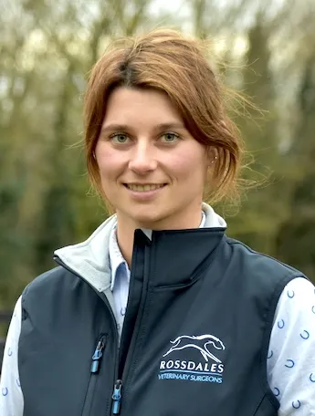 Portrait of a woman with light brown hair wearing a black vest with Rossdales Veterinary Surgeons logo, outdoors with blurred greenery in the background.