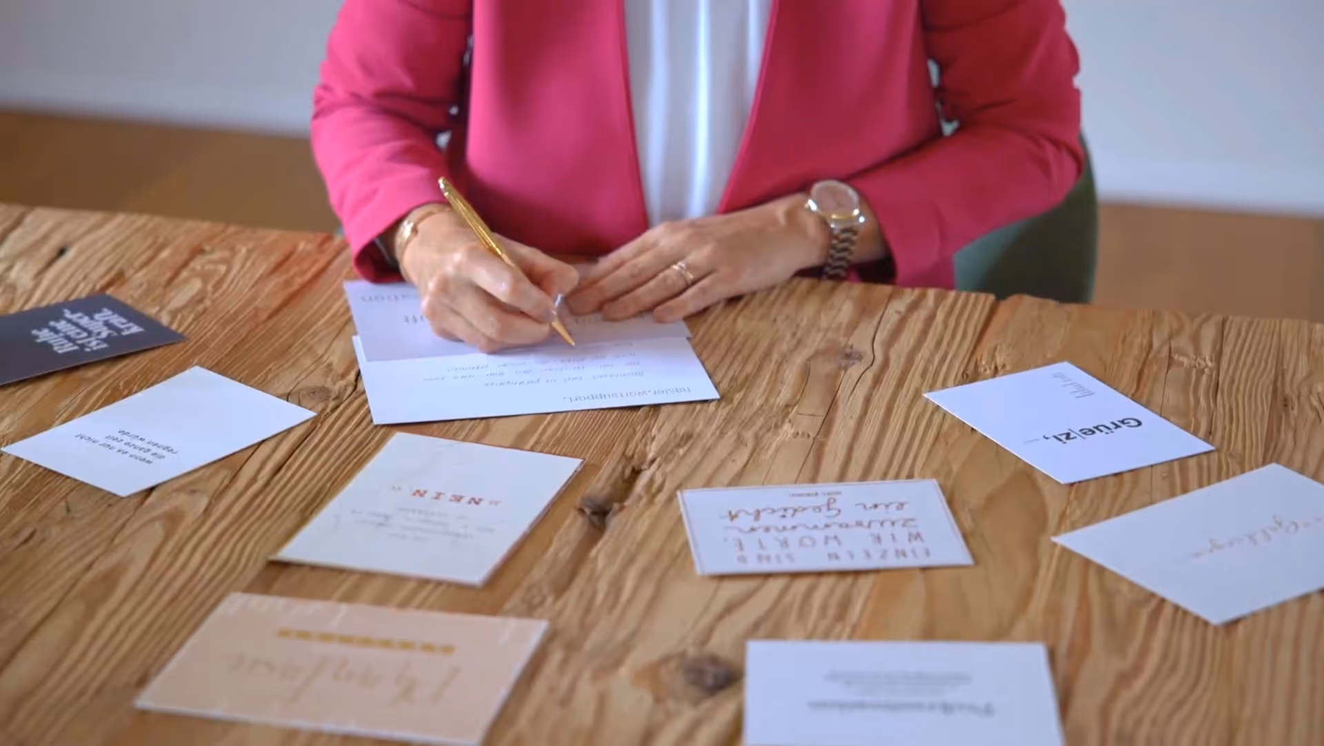 Eine Frau in pinkfarbener Jacke schreibt mit einem goldenen Stift auf eine Postkarte an einem Holztisch mit weiteren Karten.