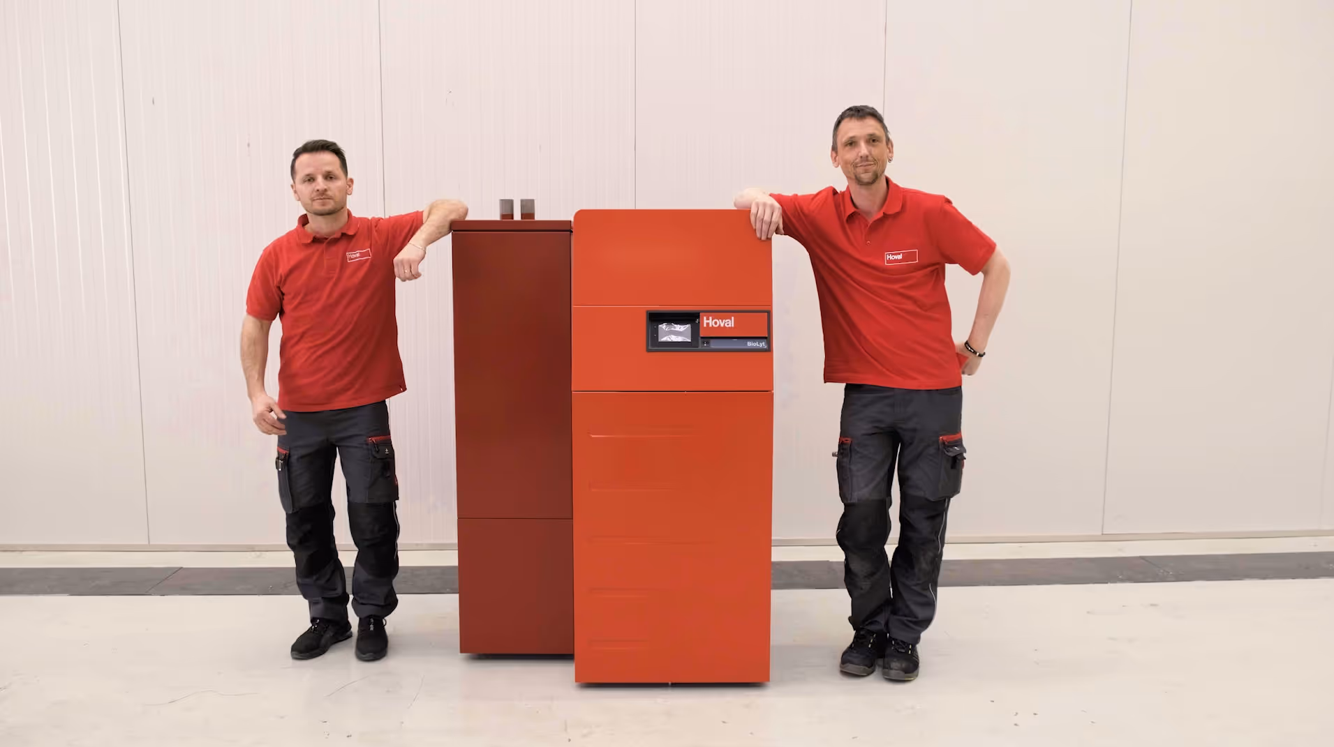 Zwei Männer in roten Hoval-Polohemden und grauen Arbeitshosen lehnen an einem roten Hoval BioLyt Heizkessel.