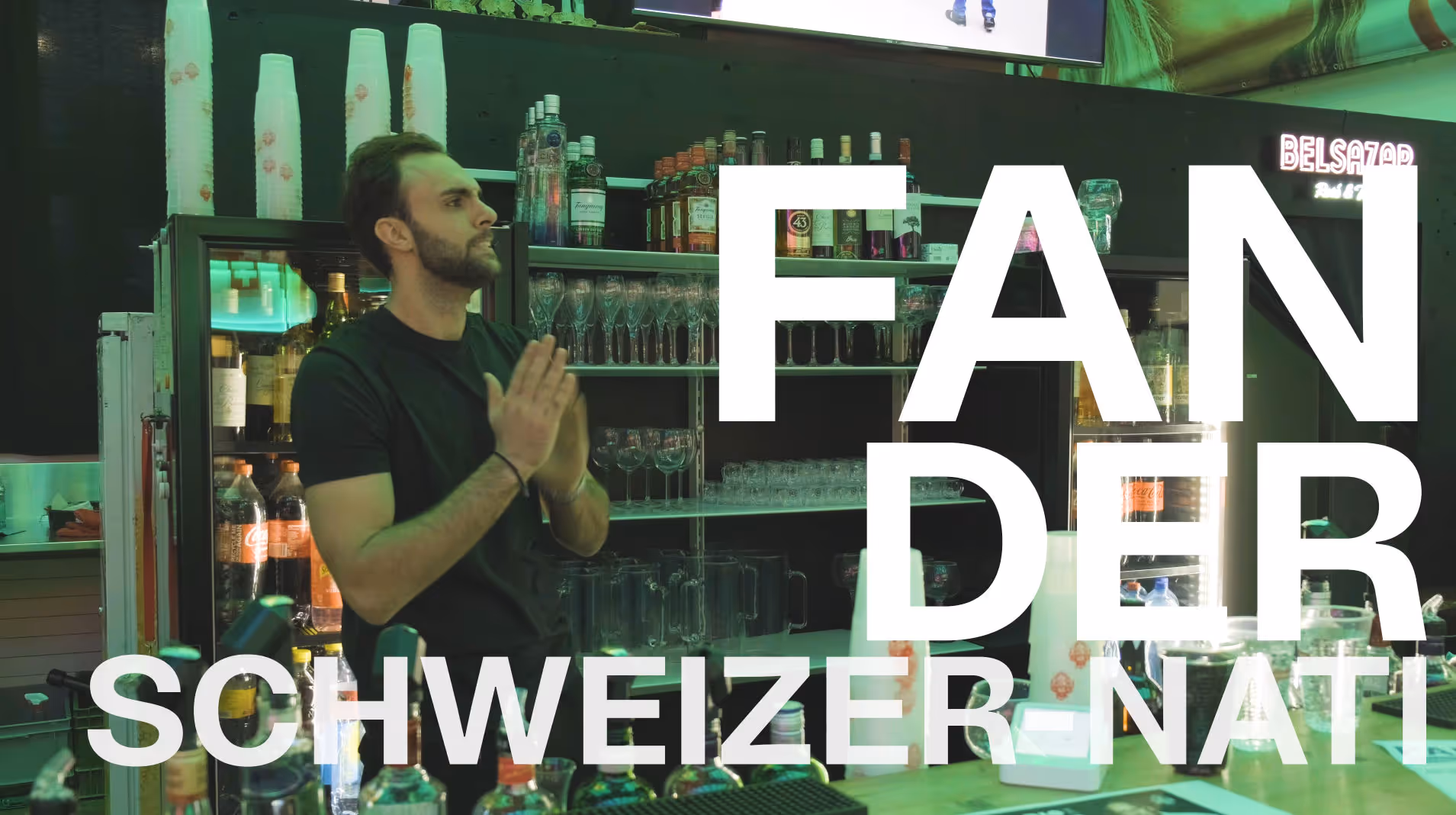 Ein Mann mit Bart hinter einer Bar mit Spirituosen, Text: "FAN DER SCHWEIZER NATI" in grosser, weißer Schrift.