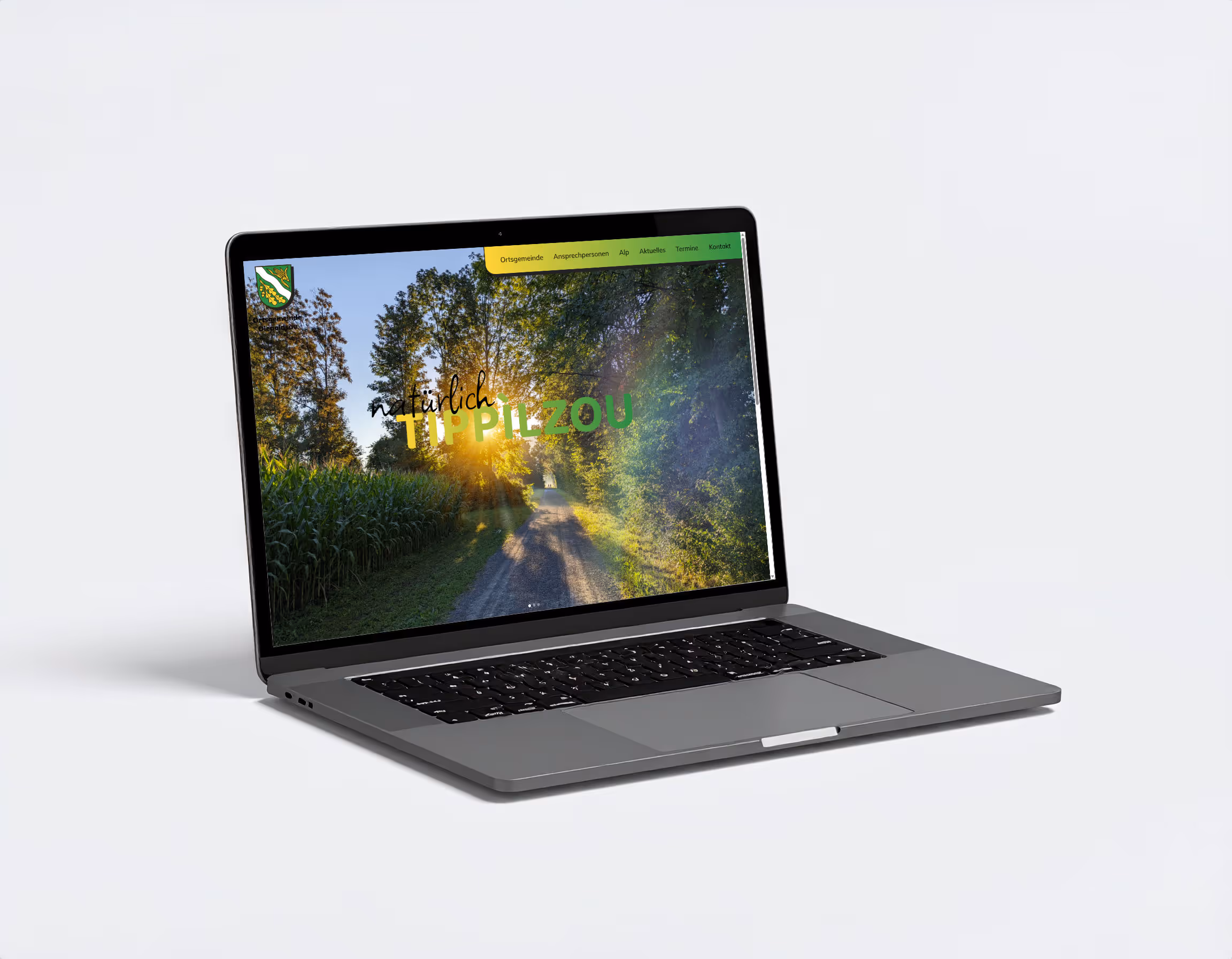 Laptop zeigt die Website der Ortsgemeinde Diepoldsau mit dem Wort "TIPPILZOU" vor einem sonnigen Feldweg.