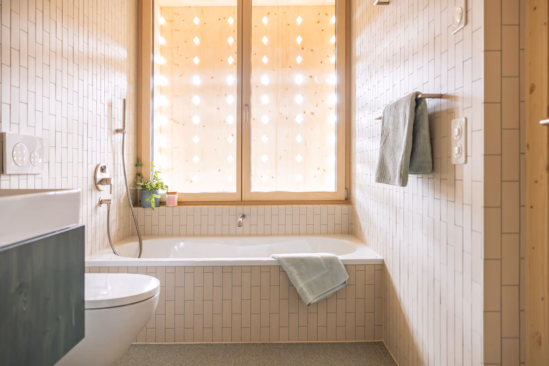 Modernes Badezimmer mit weißer, rechteckiger Badewanne, weißen Fliesen und einem Holzfenster mit Lichtpunkten.