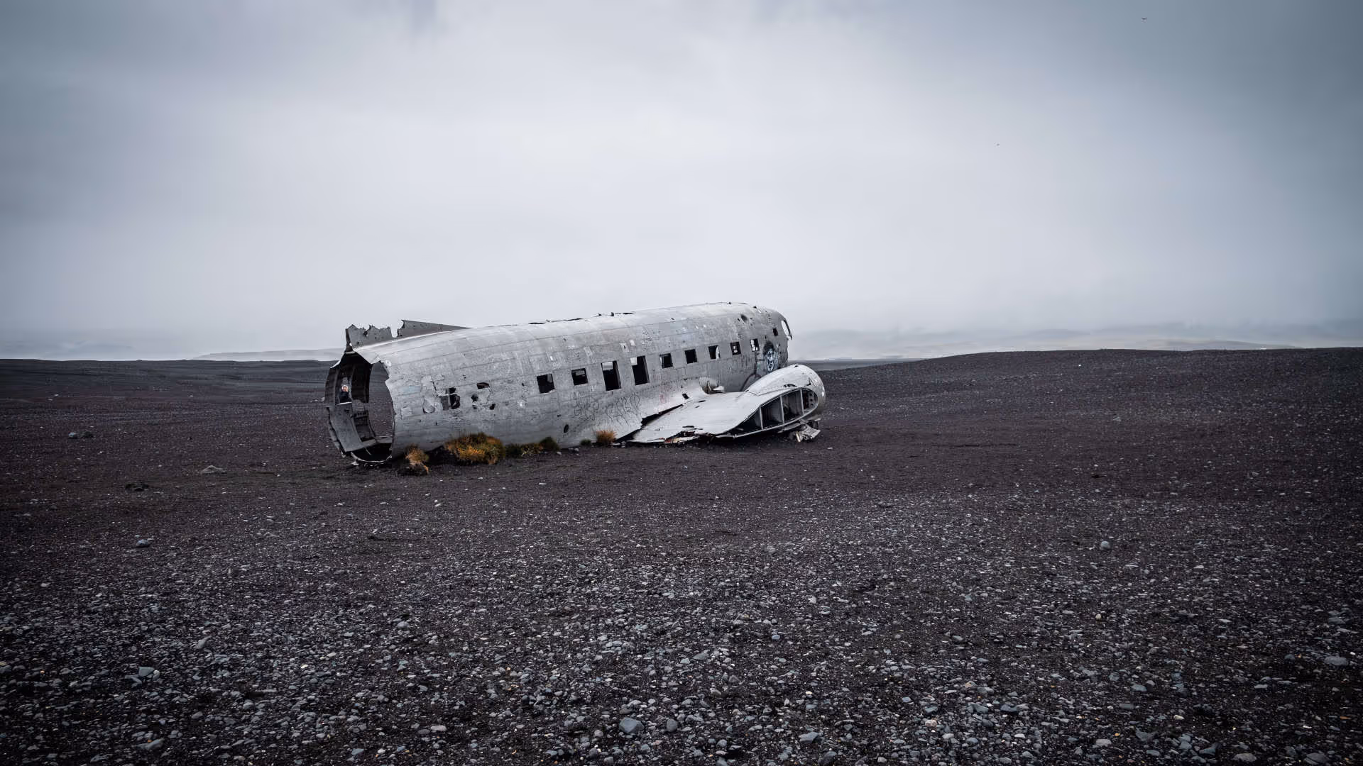 Foto vum Hutter. Das Wrack eines DC-3-Flugzeugs liegt auf einem schwarzen Kiesstrand in Sólheimasandur, Island, unter grauem Himmel.