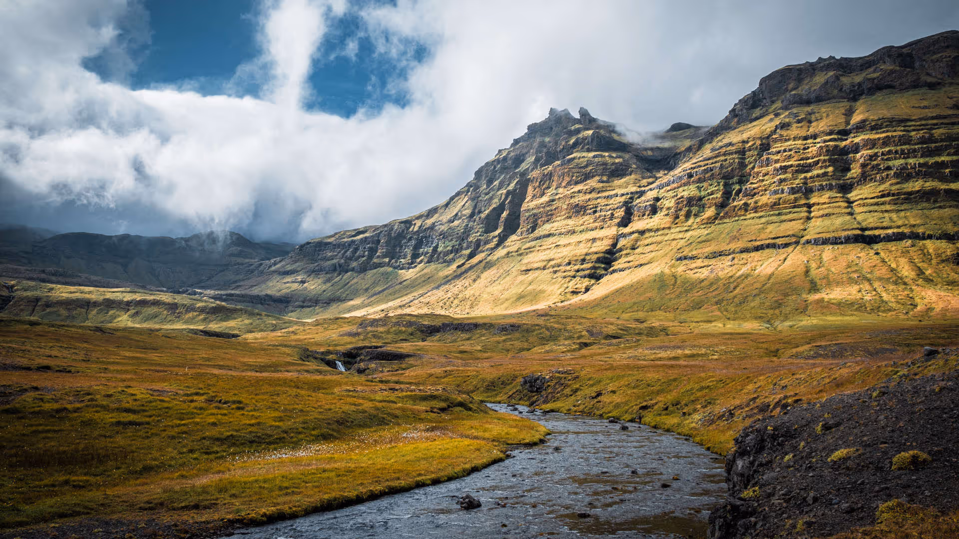 Foto vum Hutter. Landschaftsbild in der Nähe des Kirkjufellsfoss Parking Lot mit einem Fluss, der sich durch eine gold-grüne Ebene schlängelt, und steilen, terrassierten Bergen in Island.