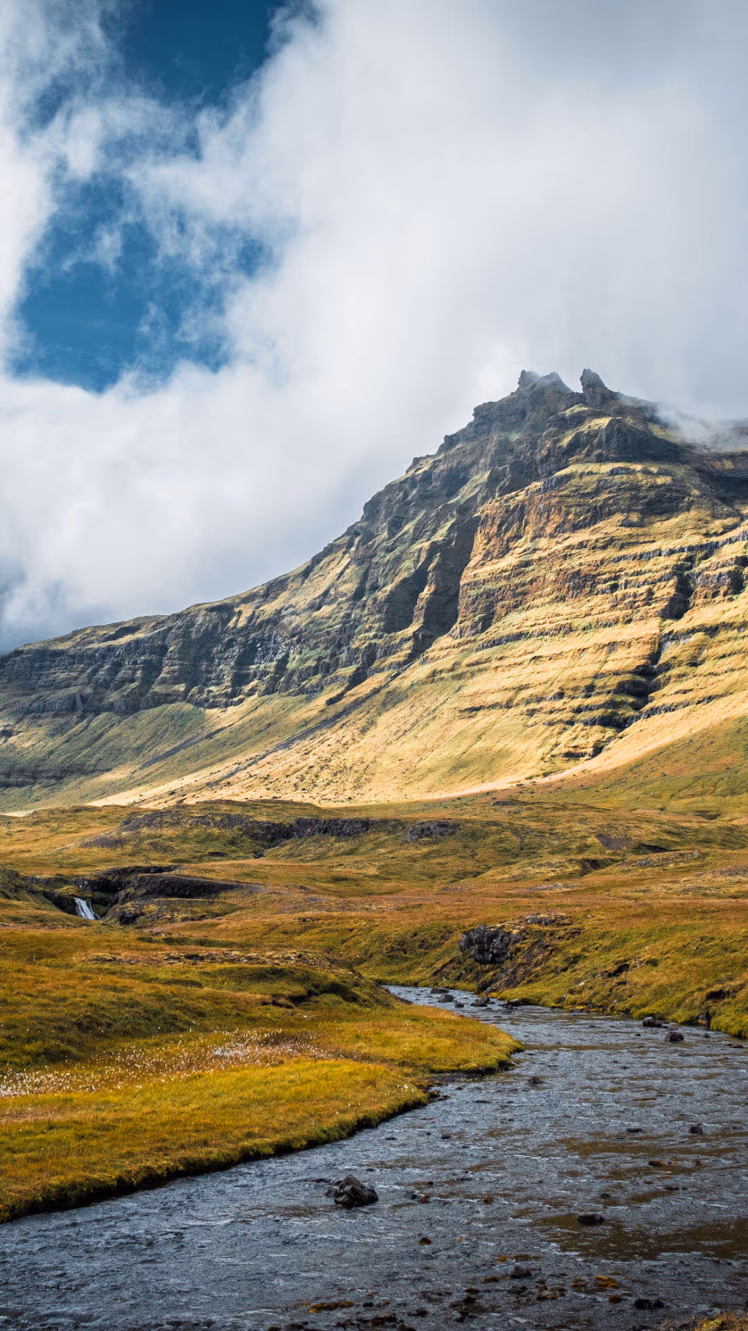 Foto vum Hutter. Landschaftsbild in der Nähe des Kirkjufellsfoss Parking Lot mit einem Fluss, der sich durch eine gold-grüne Ebene schlängelt, und steilen, terrassierten Bergen in Island.
