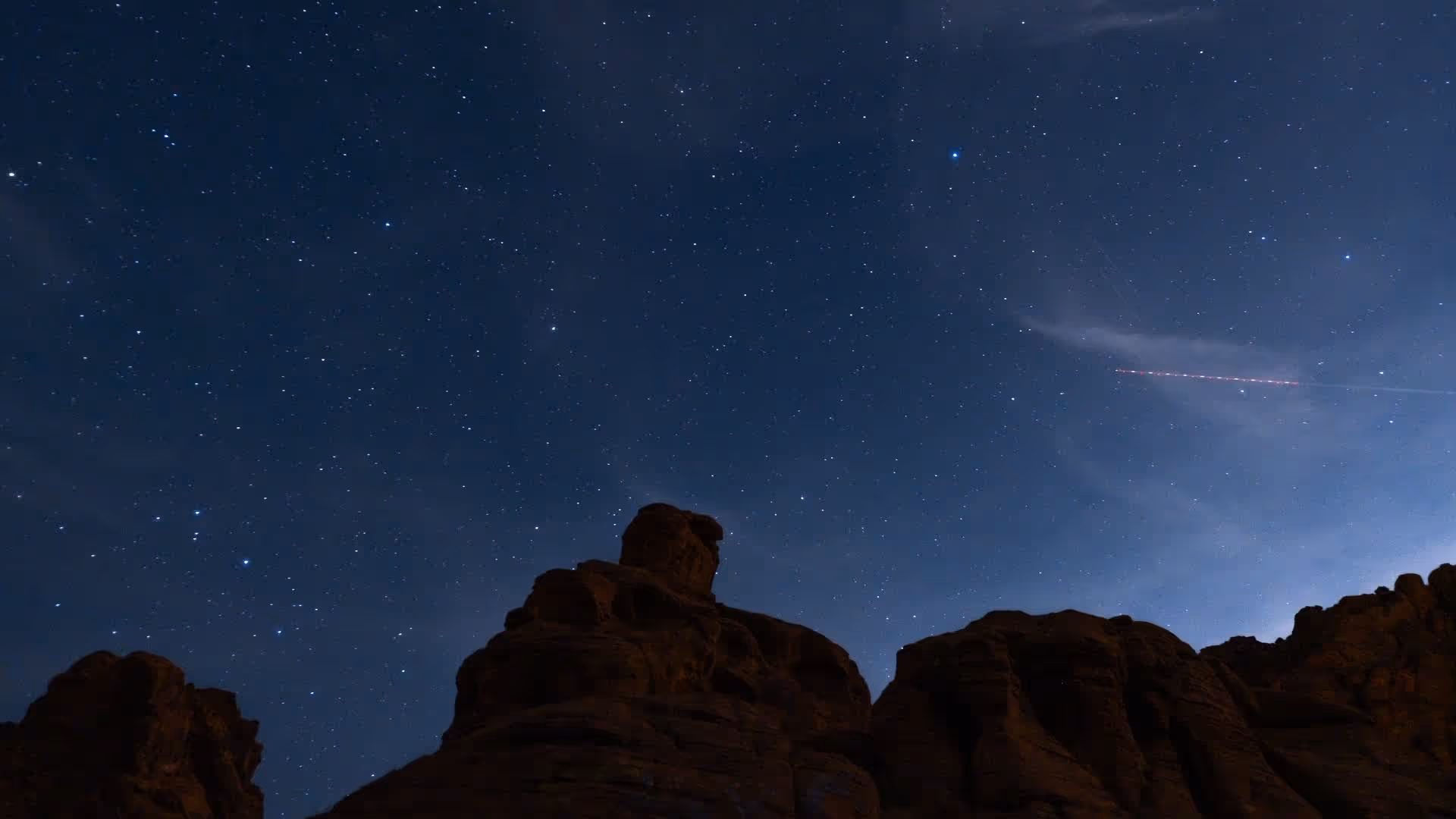 Nachtaufnahme mit sternklarem, dunkelblauem Himmel über einer dunklen Felsformation im Valley of Fire State Park; ein roter Streifen einer Sternschnuppe oder eines Flugzeugs durchquert den Himmel.