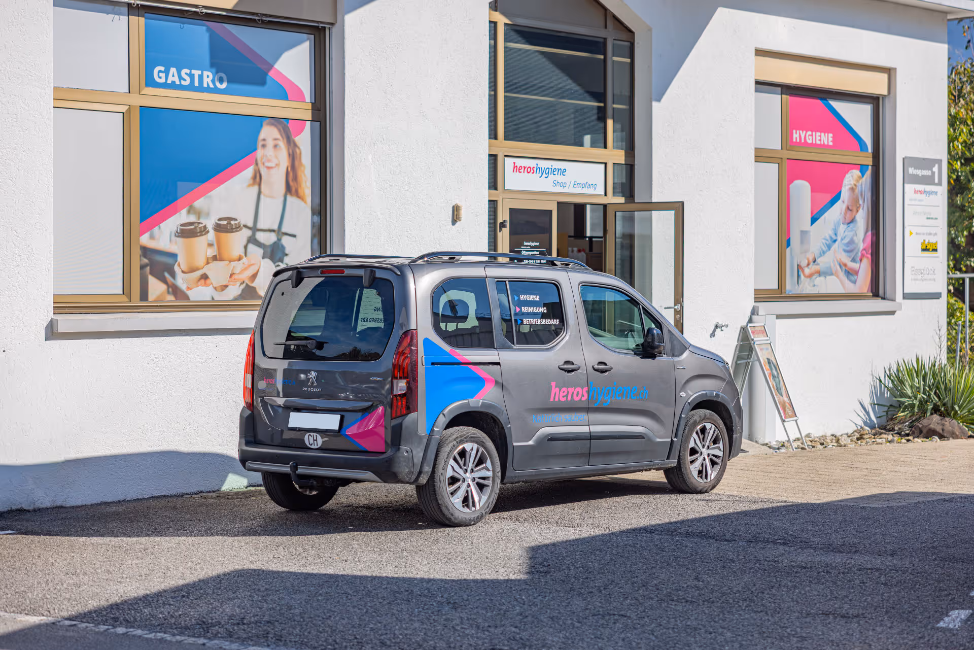 Ein gebrandeter grauer Peugeot Hochdachkombi mit pink-blauer Folierung und heroshygiene.ch Logo vor einem Geschäft.