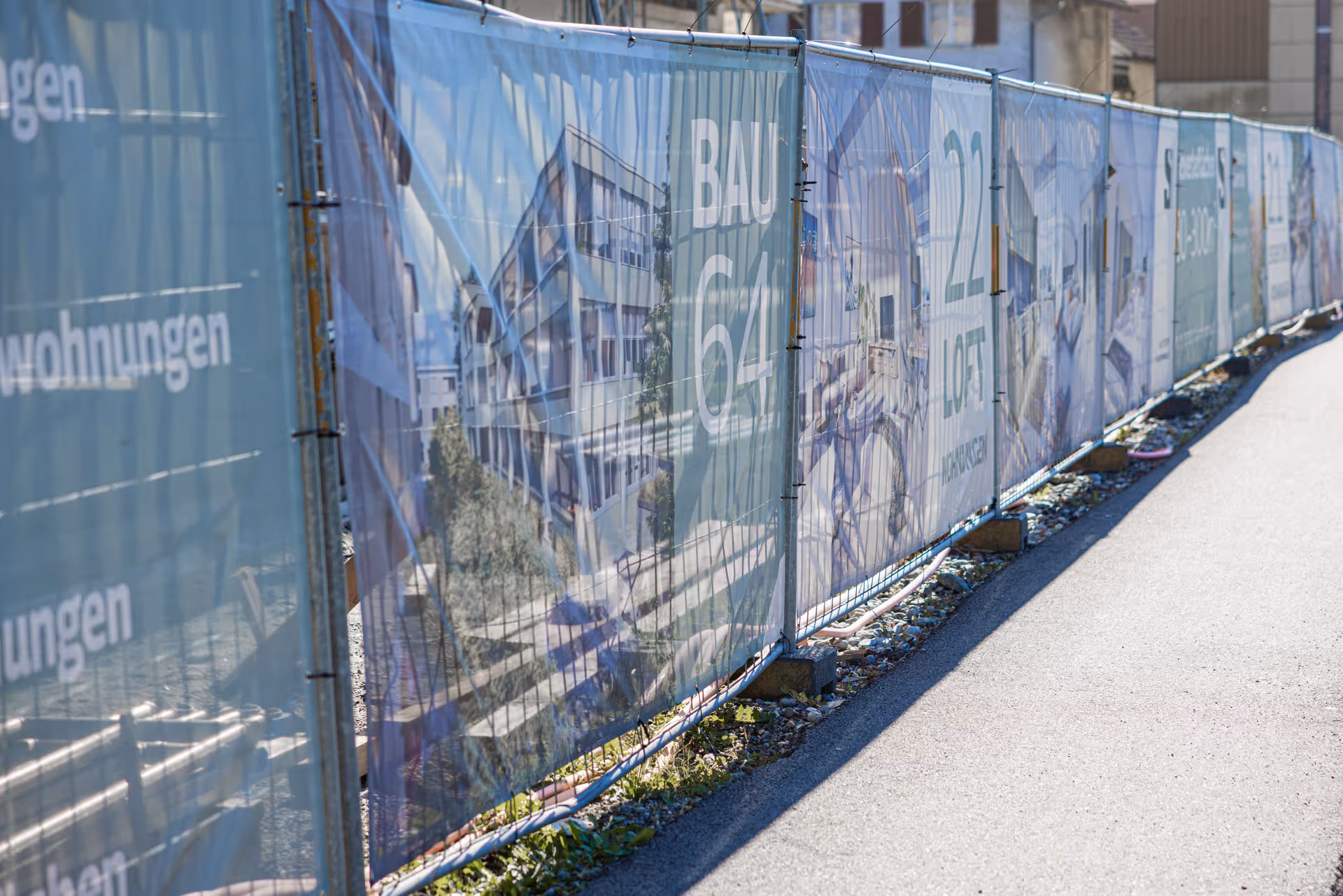 Baustellen-Sichtschutzwand mit bedruckten Werbebannern für Neubauwohnungen entlang eines Asphaltweges.