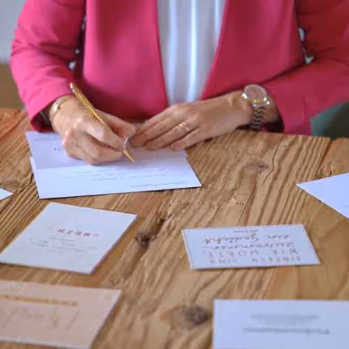 Eine Frau in pinkfarbener Jacke schreibt mit einem goldenen Stift auf eine Postkarte an einem Holztisch mit weiteren Karten.