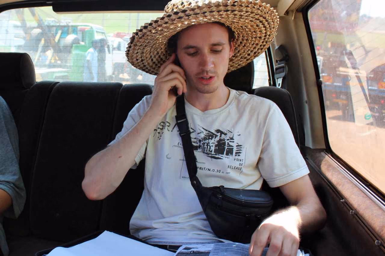 Ein junger Mann mit Strohhut telefoniert auf dem Rücksitz eines Autos und blickt auf Unterlagen in seiner Hand.