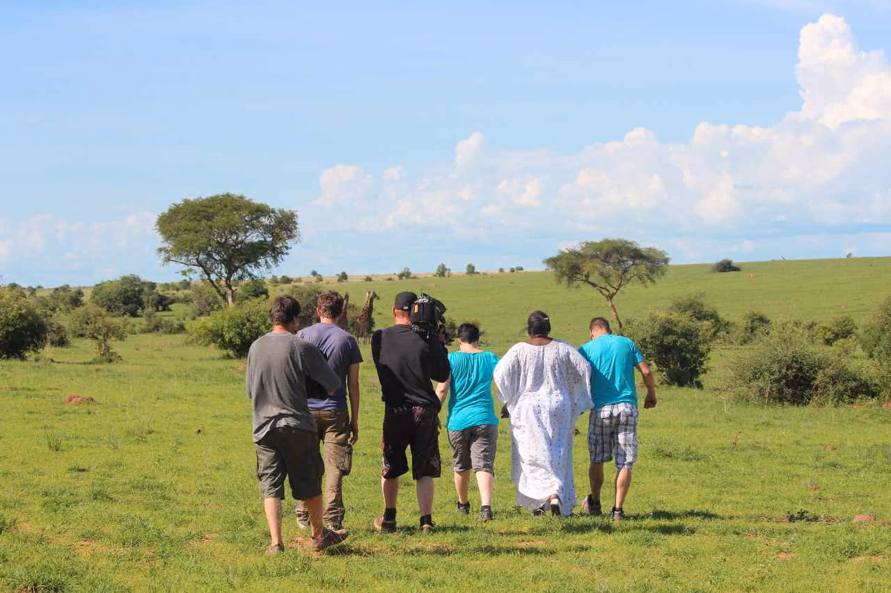 Ein Kamerateam geht zusammen mit einer Personengruppe über eine grüne Savannenlandschaft in Uganda im Freien.