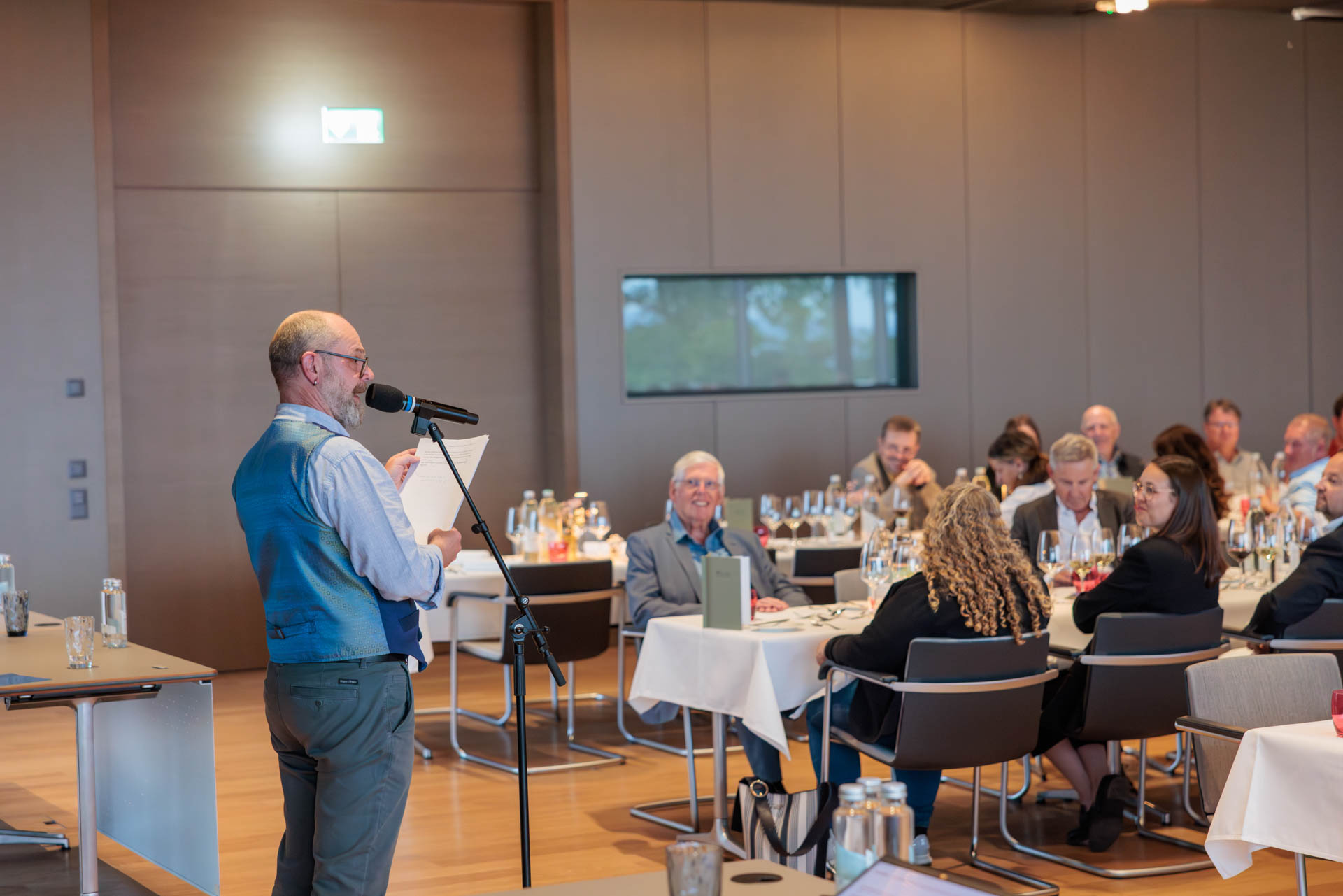Slampoet Richi Küttel am Mikrofon im Saal, Gäste an weiss gedeckten Tischen hören lachend zu
