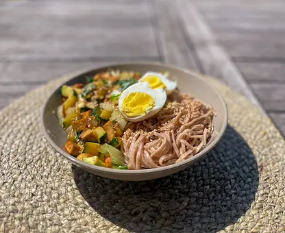 Bol de ramen aux nouilles de sarrasin avec œuf mollet coupé en deux, légumes sautés et bouillon, servi sur un set de table en osier