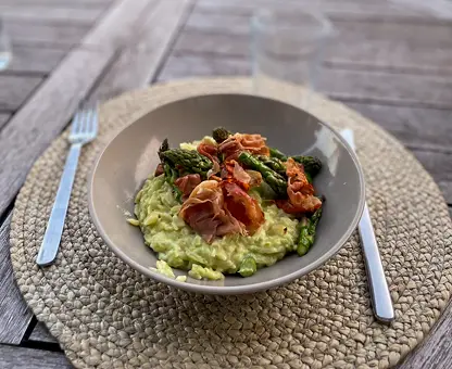 Assiette de risotto crémeux aux asperges vertes, garnie de tranches de jambon croustillant, posée sur un set de table en fibres naturelles avec des couverts de chaque côté.