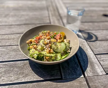 Bol de salade estivale sur table en bois avec lamelles de concombre, graines, herbes fraîches et vinaigrette, accompagné d'un verre d'eau