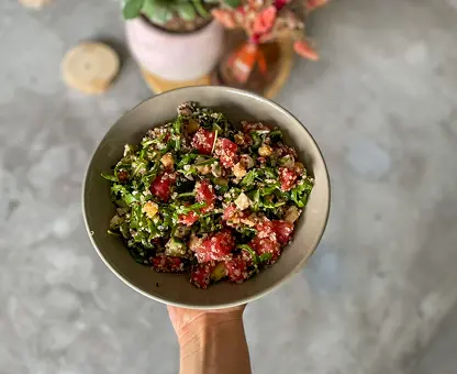 Bol de salade colorée tenu à la main, composée de quinoa, légumes verts feuillus, tomates cerises, graines et autres ingrédients variés, présentée sur un plan de travail en marbre avec des éléments de cuisine en arrière-plan