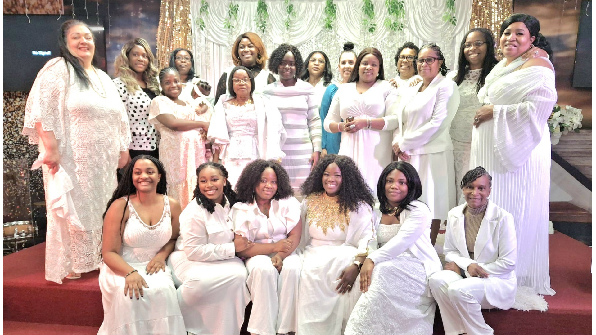 Group of diverse women and girls dressed mostly in white, posing on red steps with decorative white and green backdrop.