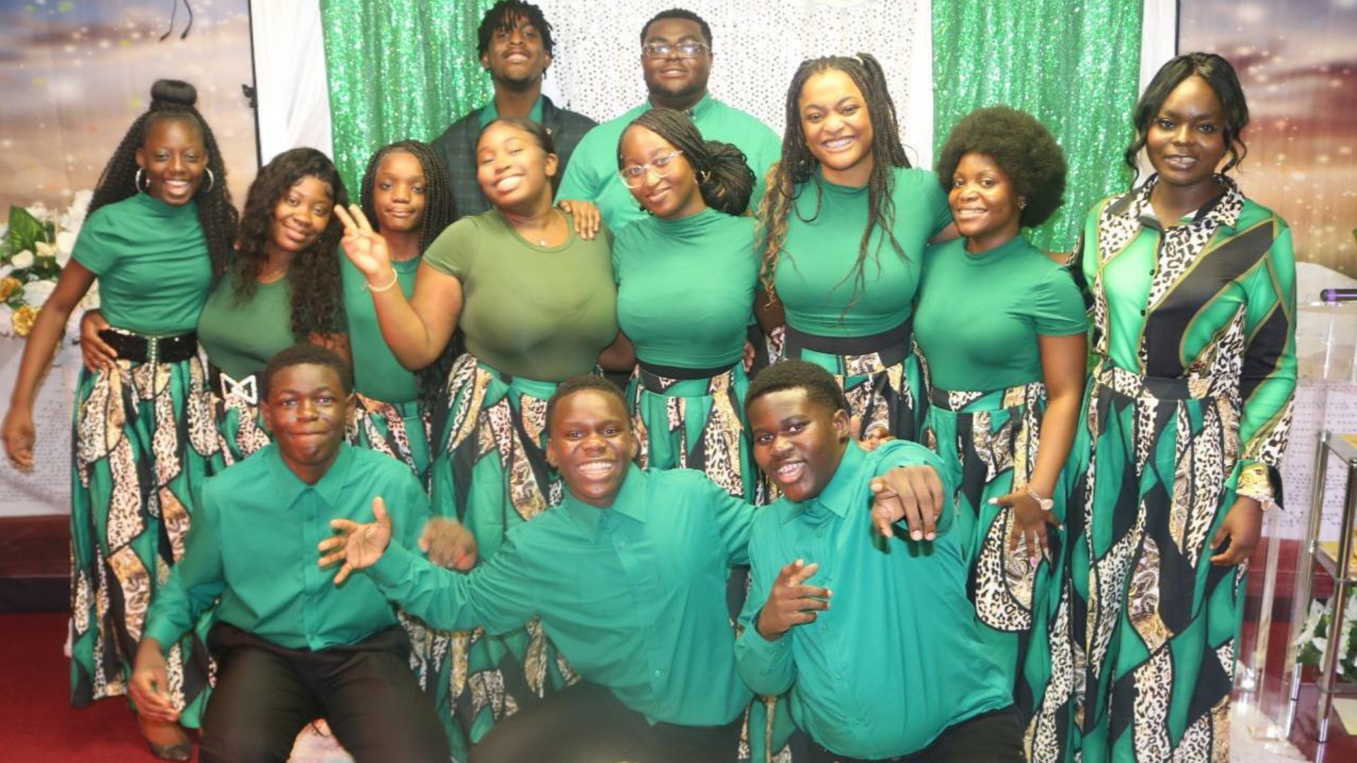 Group portrait of smiling young men and women wearing coordinated green tops and patterned skirts or pants, posing indoors with green and white sparkling backdrop.