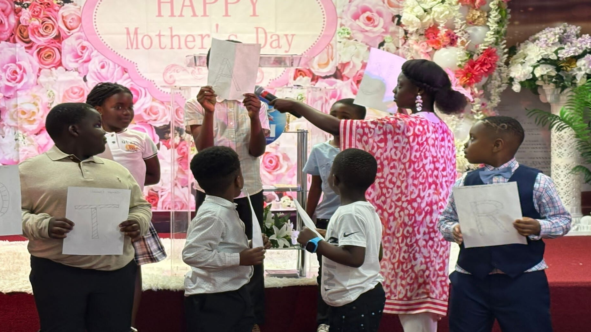 A group of children holding papers with letters during a Mother's Day event with floral decorations and a woman holding a microphone.