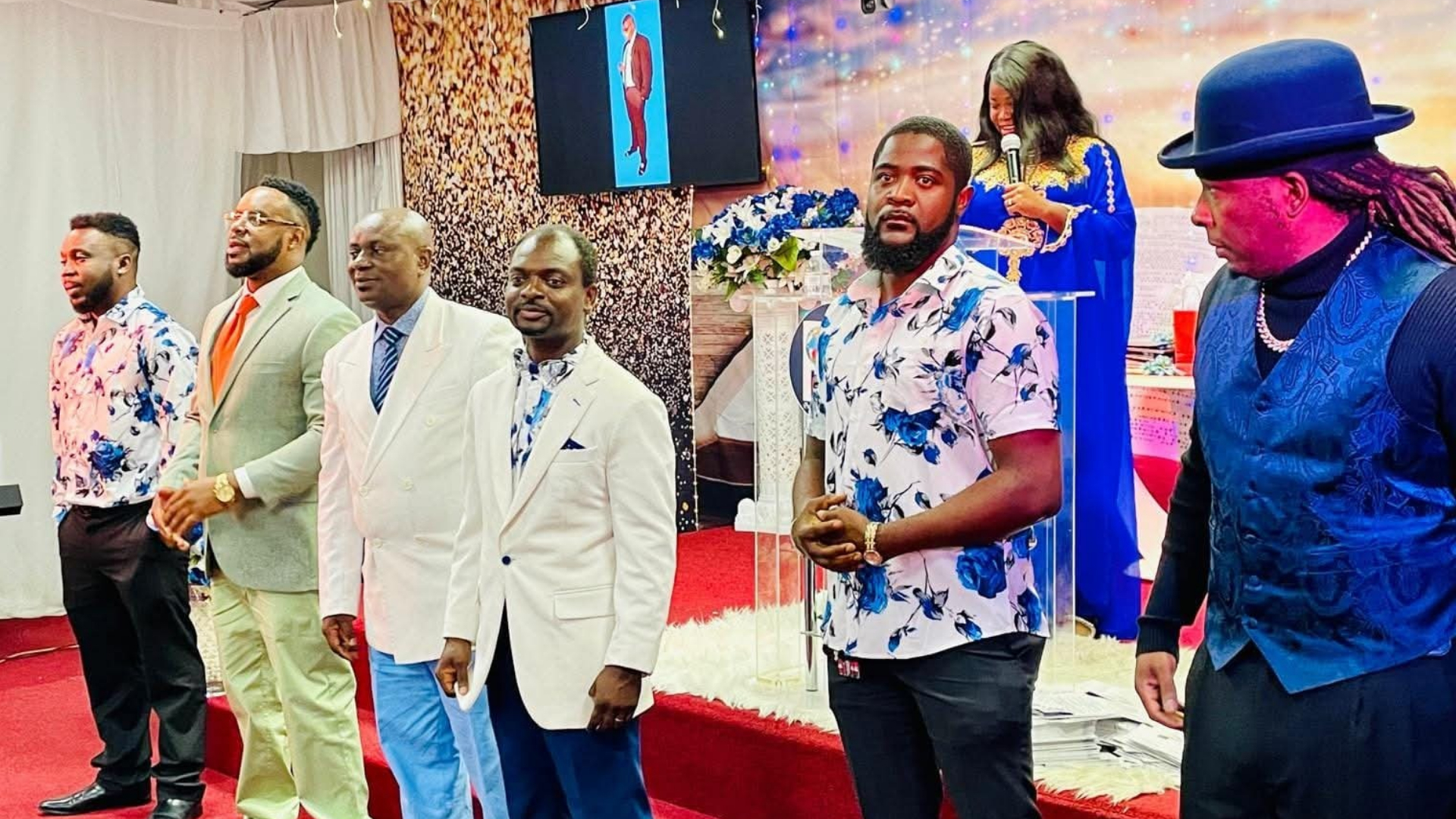 Six men standing in a row inside a decorated room with a woman speaking into a microphone in the background.