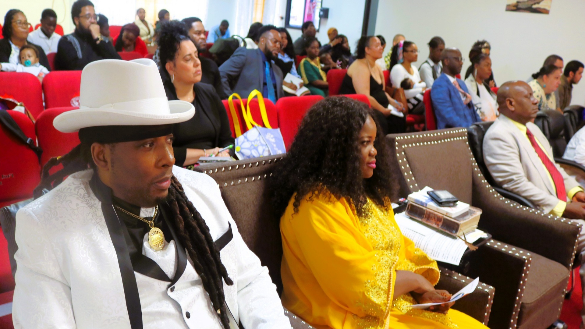 Audience seated in a room with a man in white suit and hat and a woman in a yellow dress in the front row.