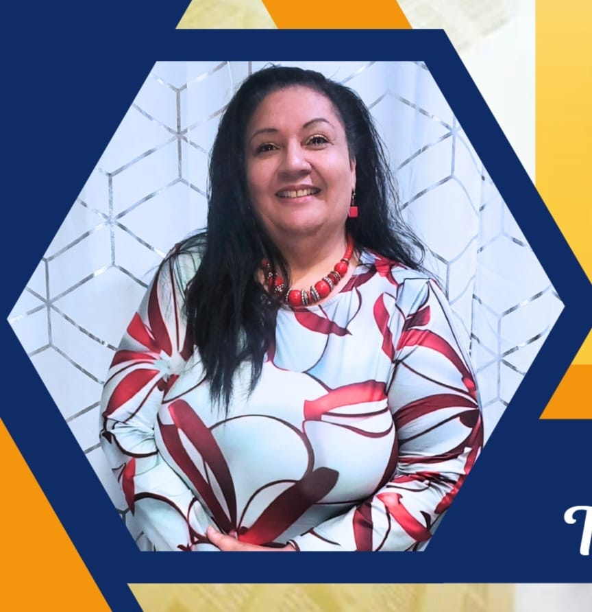 Woman with long dark hair wearing a white top with red floral patterns and red beaded necklace, smiling in front of a geometric-patterned background.