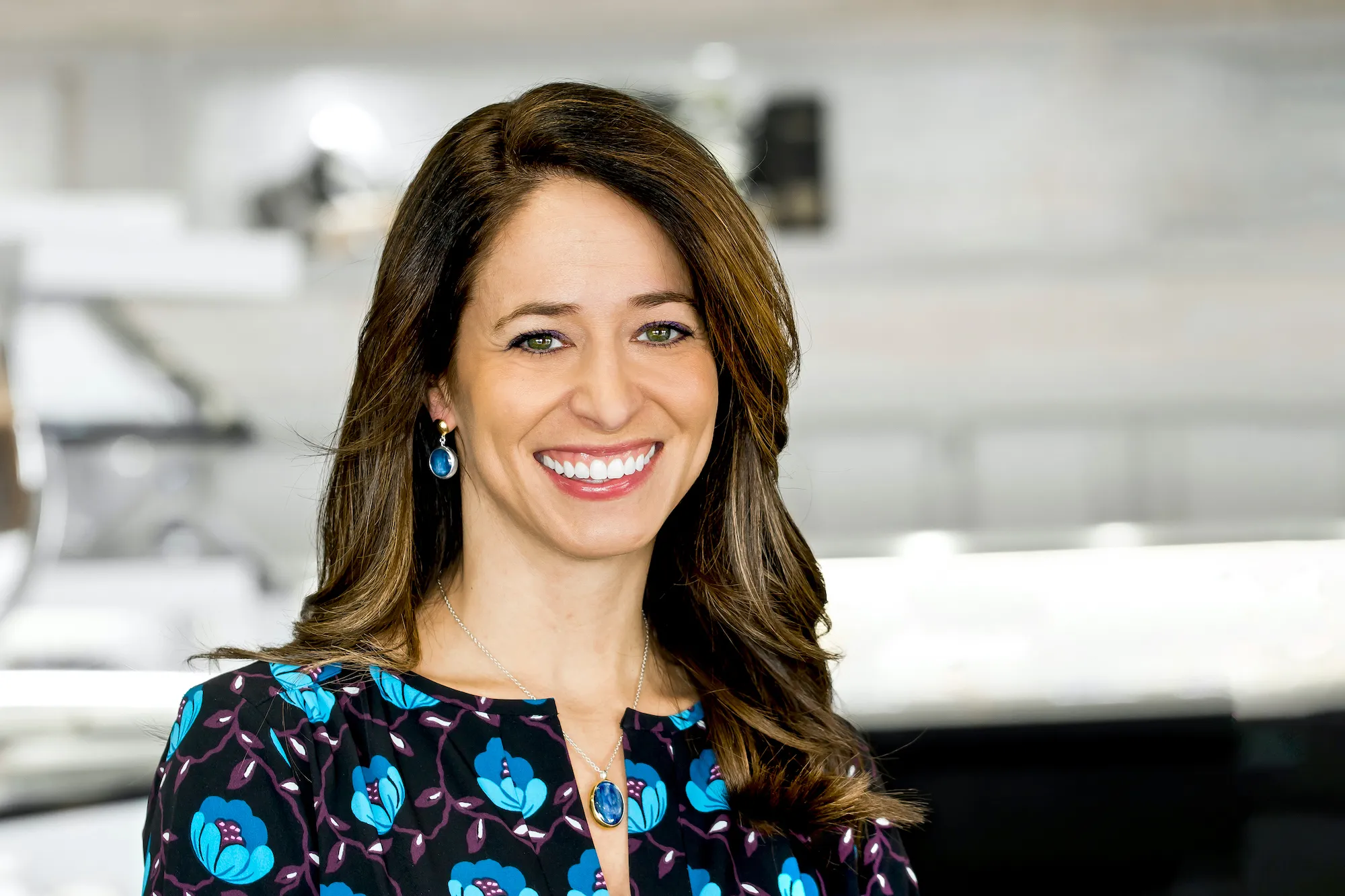 Headshot of Sales Manager Missy Ranney. With long brown hair and a big smile she is looking at the camera wearing a shirt with blue flowers and blue tear drop earrings.