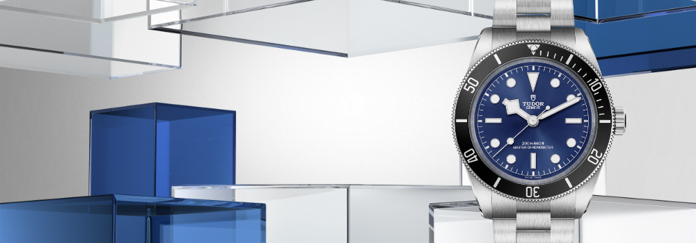 The Black Bay 68 features a 43mm stainless steel case with polished and satin finishes, a sun-brushed “TUDOR Blue” dial, and a 60-minute unidirectional bezel in stainless steel with matte black aluminum insert. Model number m7943a1a0nu-0001 appears on the right side of the screen on a modern silver and blue geometric background. Jack Lewis Jewelers located at 3011 Galena Rd, Bloomington, Illinois, is proud to be part of the worldwide network of Official Tudor Jewelers, allowed to sell and maintain Tudor Watches.