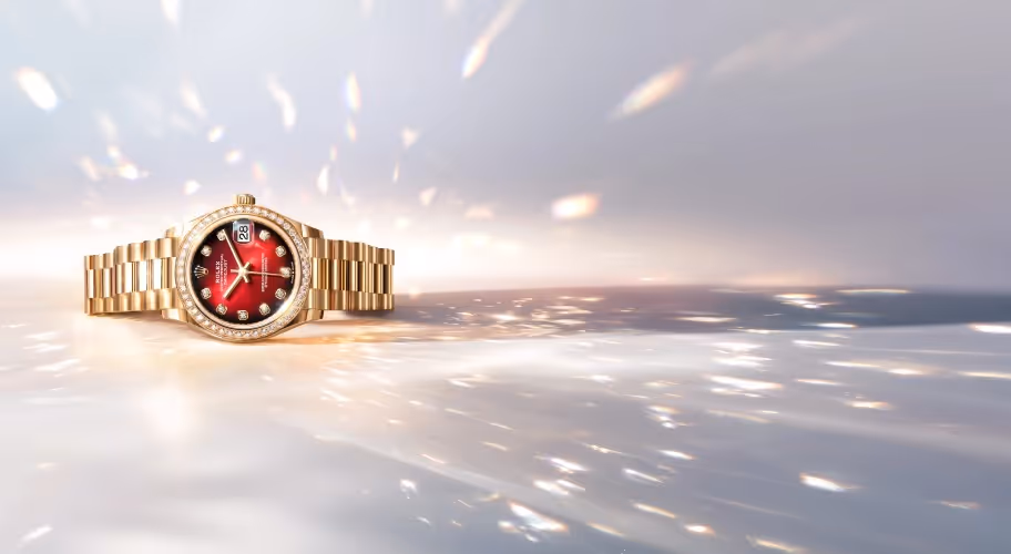 Positioned to the left against a luminous, shimmering backdrop, the Rolex Datejust in yellow gold features a striking red dial illuminated by diamond hour markers. Its elegant profile is enhanced by a diamond-set bezel and the iconic President bracelet, reflecting light in a cascade of warm gold tones. Equipped with the signature date display at 3 o’clock and crafted for precision, reliability, and enduring style, this Datejust embodies timeless luxury. Jack Lewis Jewelers of Bloomington, Illinois, now at 3011 Galena Rd, is proud to be part of the worldwide network of Official Rolex Jewelers.