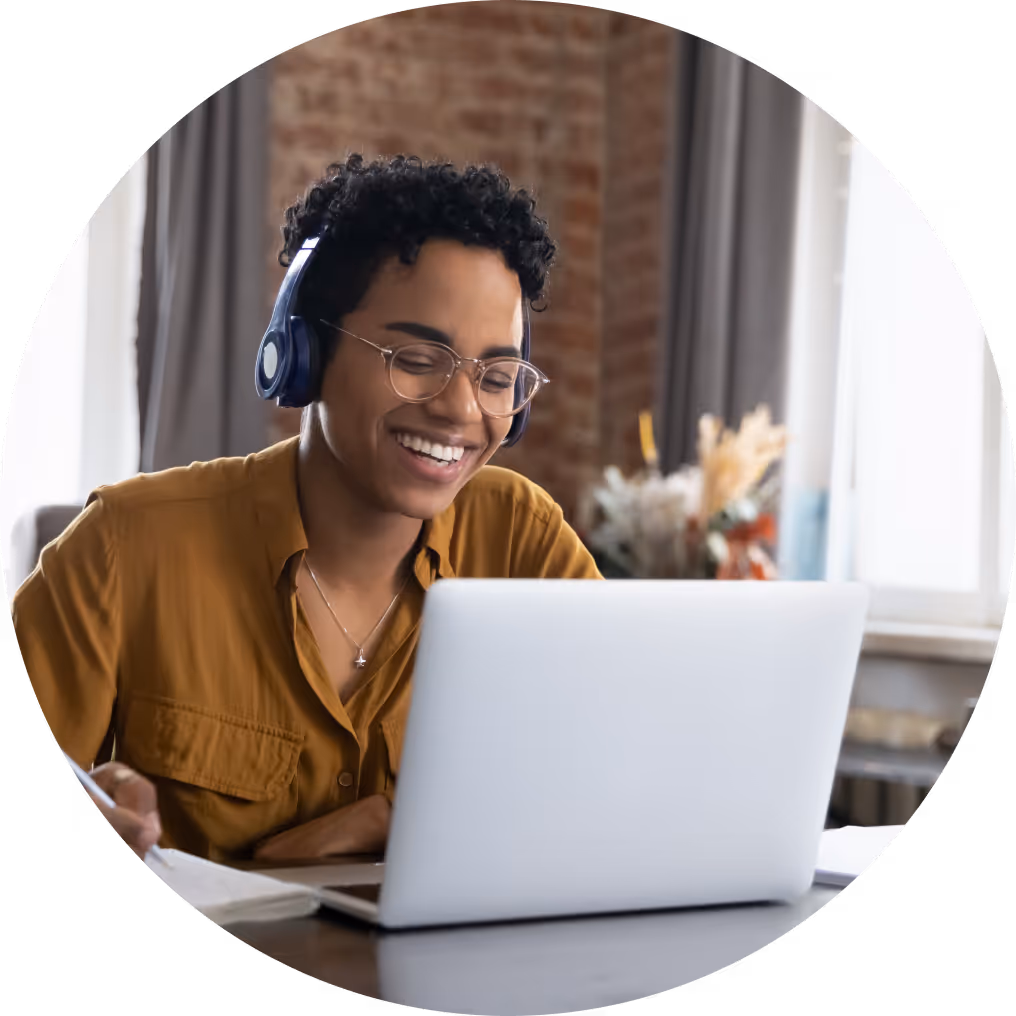 Woman wearing headphones smiling while working on laptop and taking notes