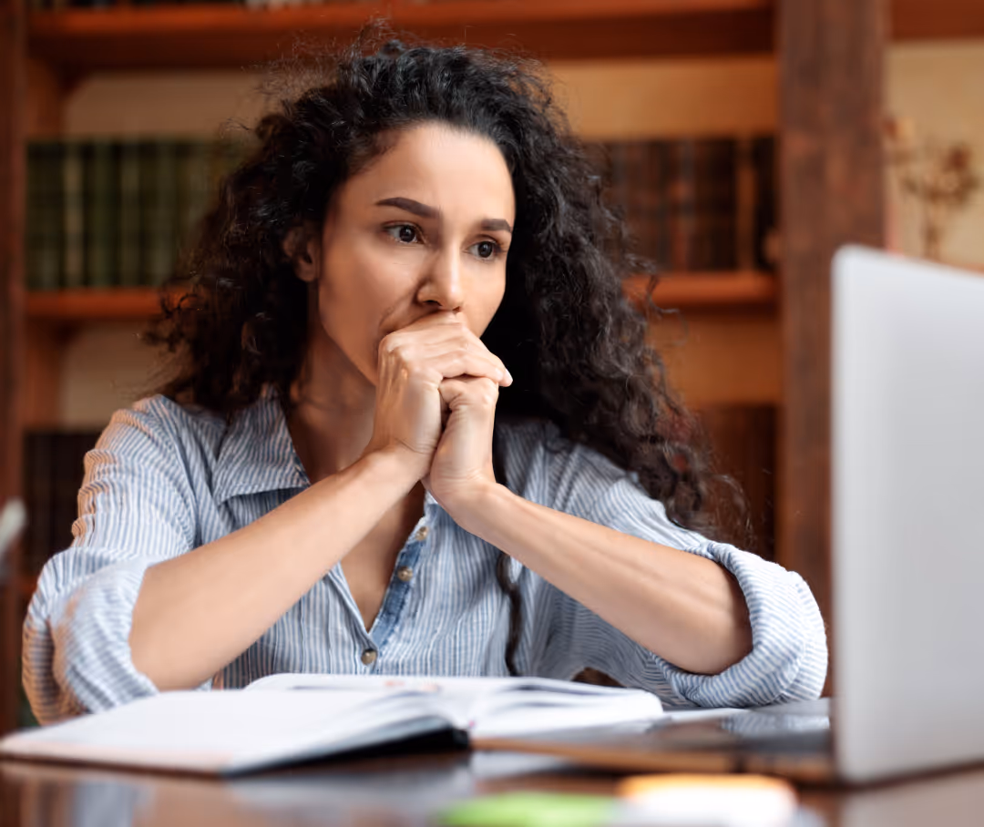A woman sitting at a desk looking concerned while watching her laptop, with an open book in front of her.
