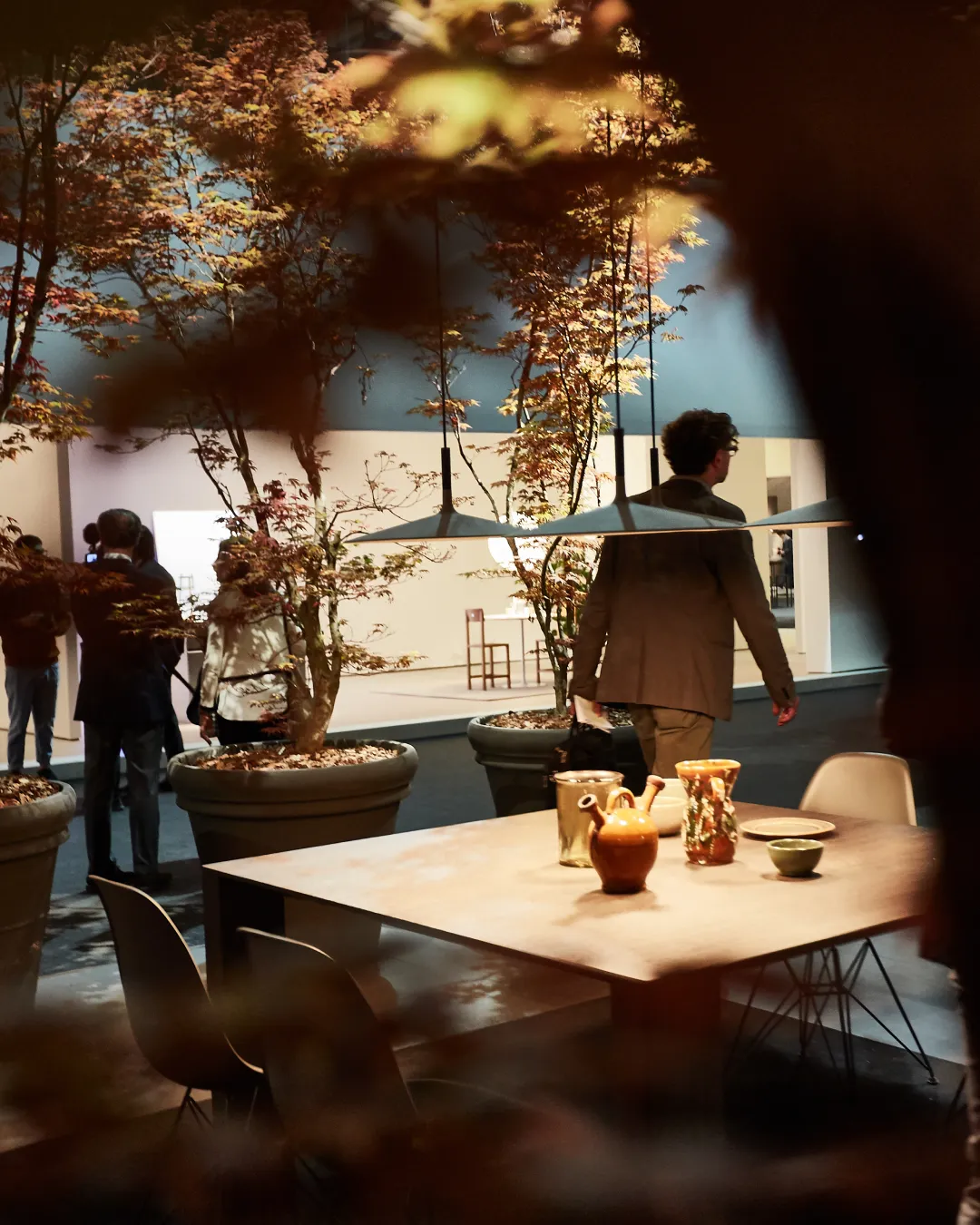 Messestand Innenansicht mit modernem Esstisch und Leuchte, umgeben von herbstlich gefärbten Topf-Ahornbäumen.