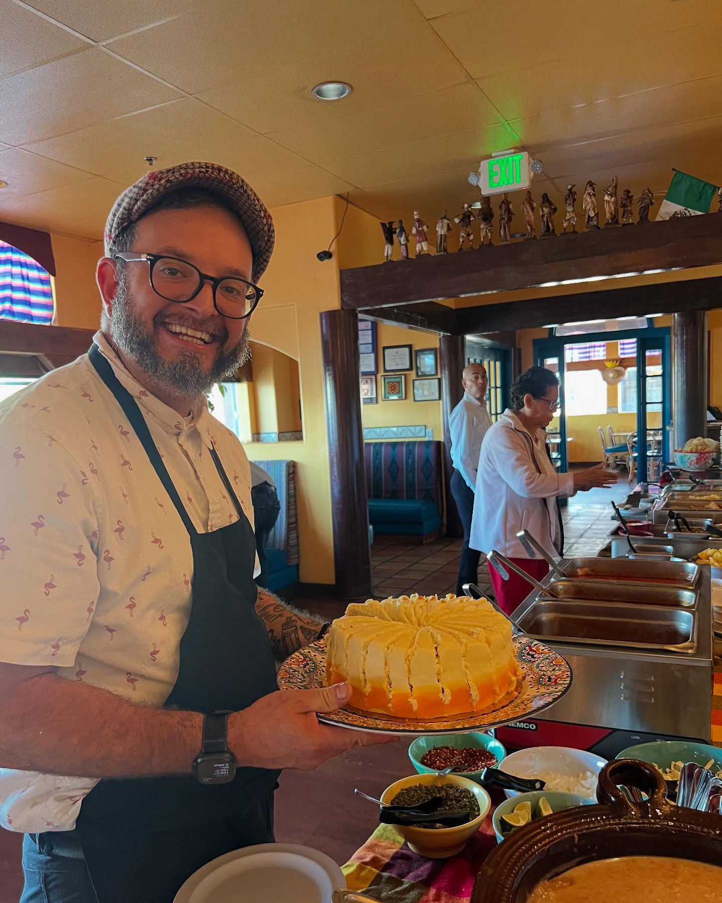 A man smiling in a restaurant, holding a cake on a decorated plate. He wears a cap and an apron. Food trays and bowls are visible on the counter.