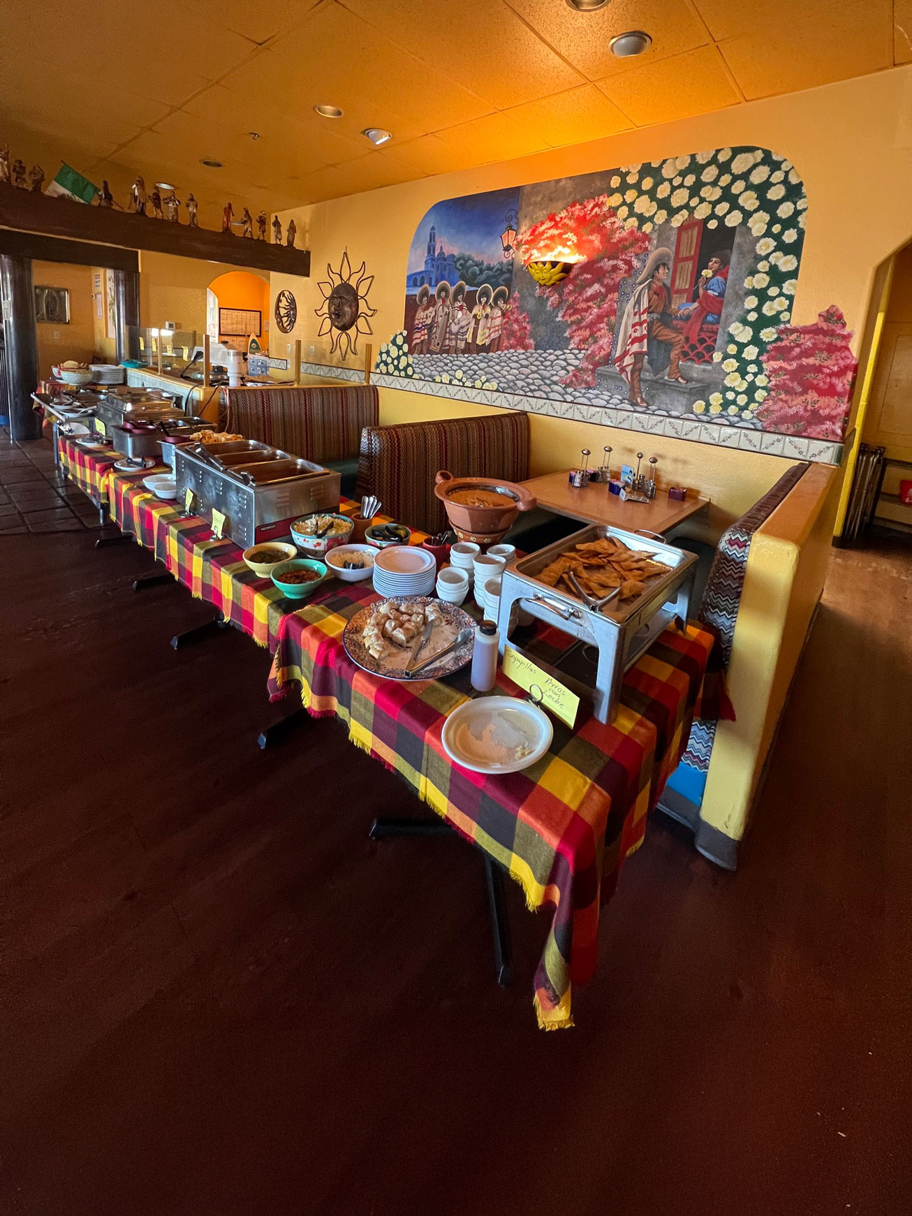 Buffet setup in a cozy restaurant with a vibrant mural. Tables are covered with colorful checkered cloths, showcasing various dishes.