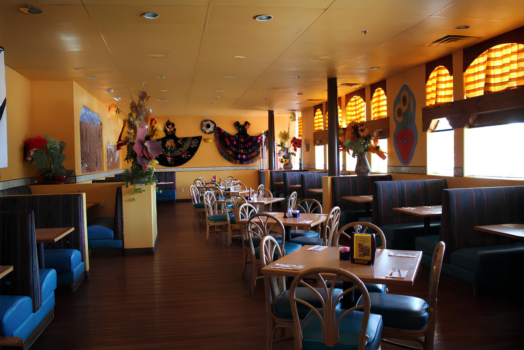 A Mexican restaurant interior with yellow walls, decorative dresses, colorful artwork, and neatly arranged tables and chairs.