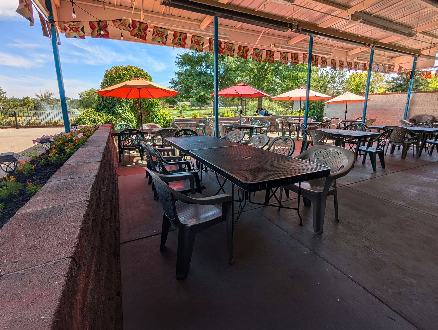 Outdoor patio with plastic chairs and tables under a covered area. Red umbrellas and lush greenery create a relaxed ambiance.
