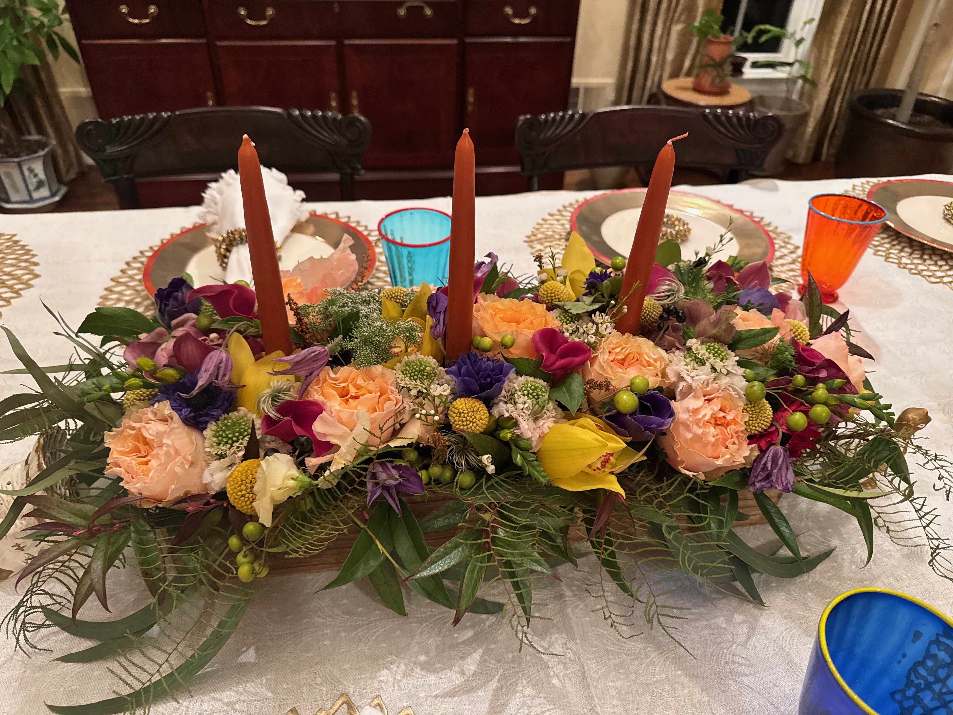 A large arrangement of flowers including peach roses, purple flowers and other green filling leaves with three red candles on a dinner table.