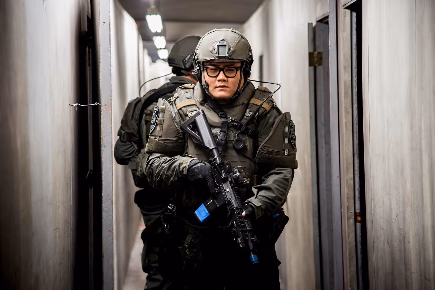 Two armed tactical officers in protective gear moving through a narrow, dimly lit corridor.