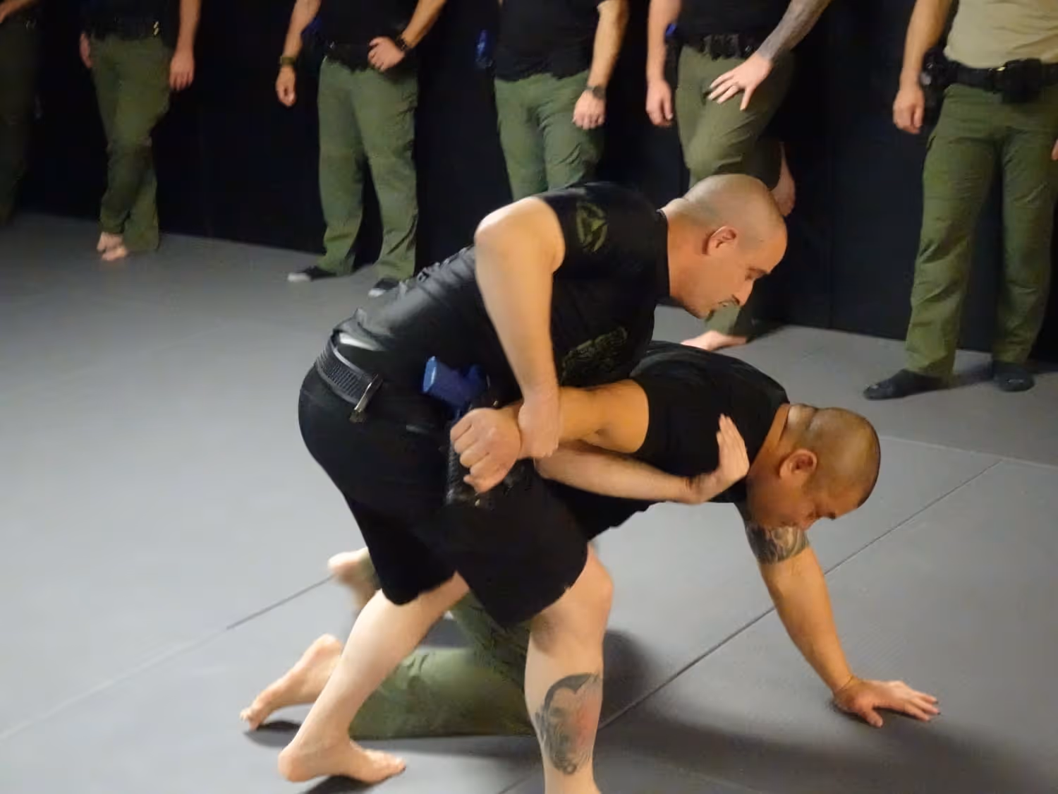 Two men practicing a grappling technique on a mat with others standing and watching in the background.