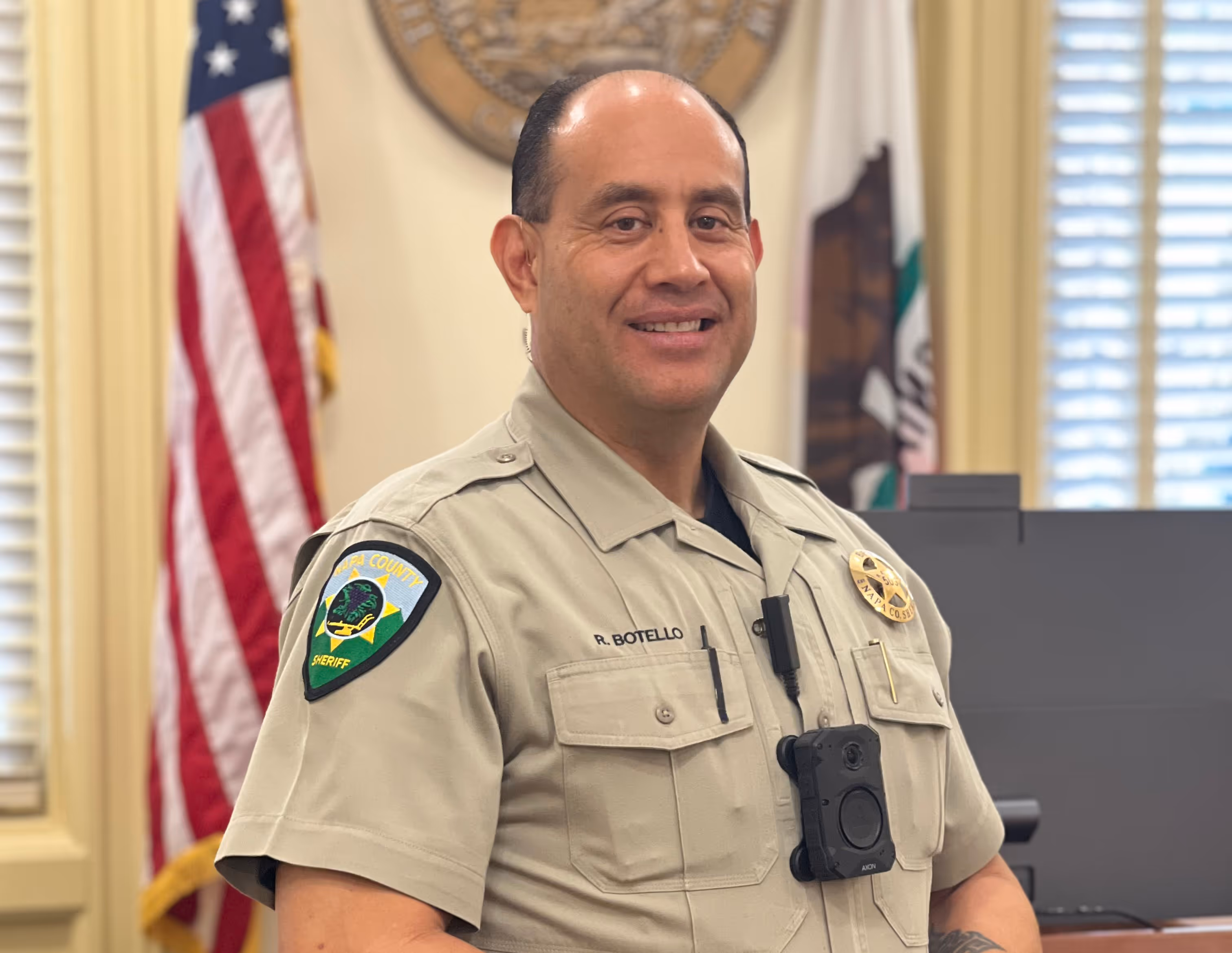Smiling sheriff officer in uniform with body camera, standing in office with U.S. and California flags behind him.