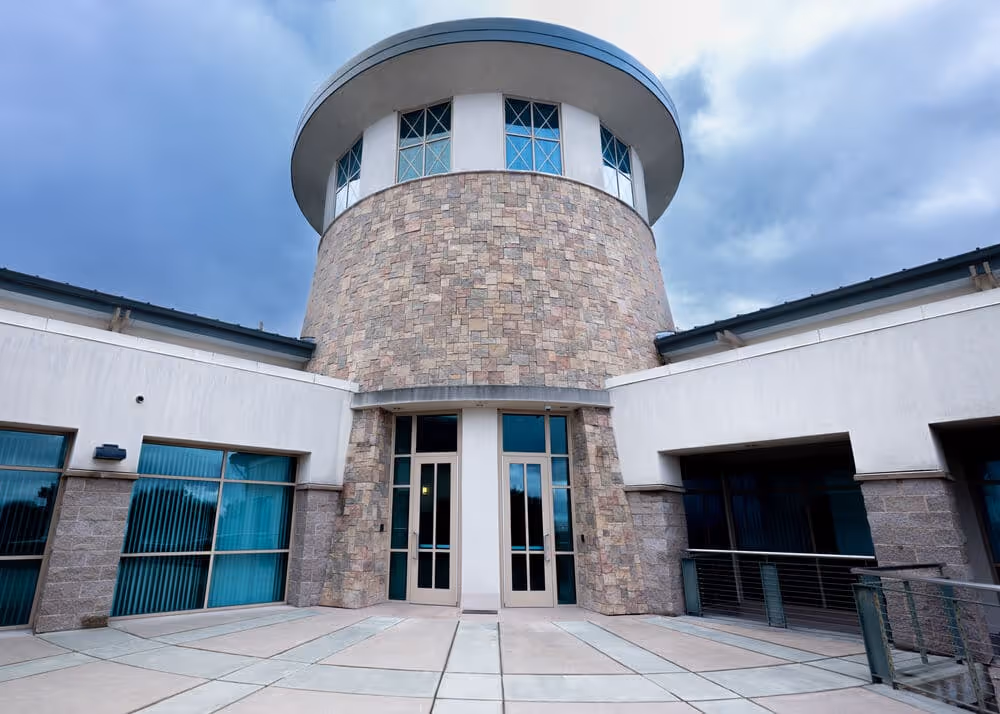 Modern building entrance with a cylindrical stone tower and glass doors under a cloudy sky.