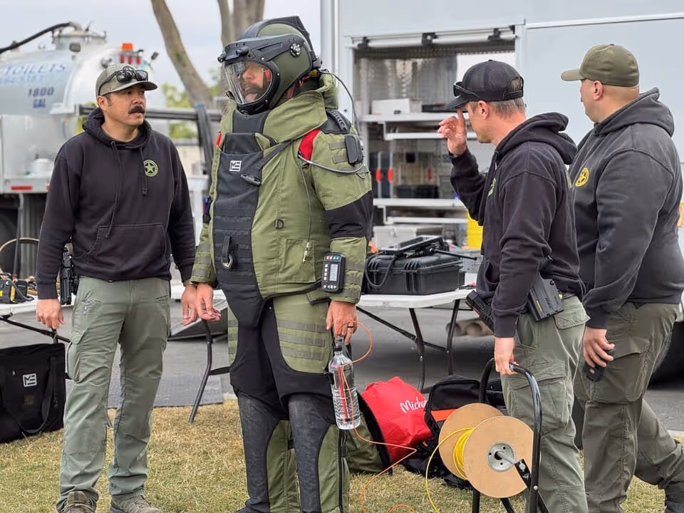 Four men, one wearing a bomb disposal suit, standing outdoors next to equipment and a white truck.