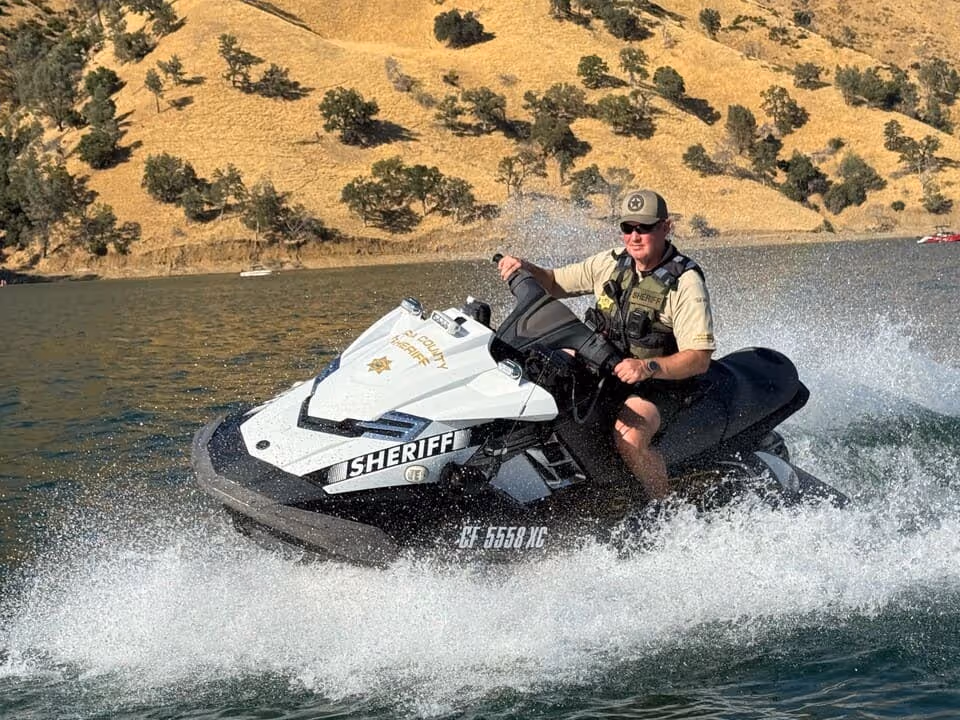 Sheriff riding a white jet ski on a lake with dry, tree-dotted hills in the background.